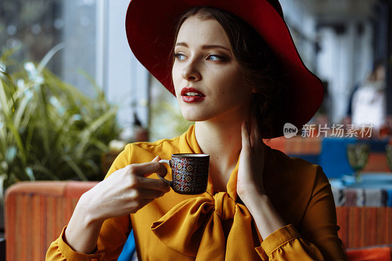 女孩在咖啡馆喝着咖啡，戴着帽子。性感的年轻女孩的肖像戴着松软的帽子和衬衫与蝴蝶结。美丽的黑发女人在咖啡馆拿着一杯咖啡