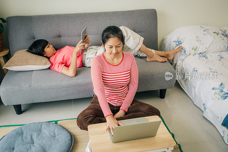 亚洲母亲坐在地板上使用笔记本电脑，她十几岁的女儿躺在家里或公寓的沙发上使用数字平板电脑