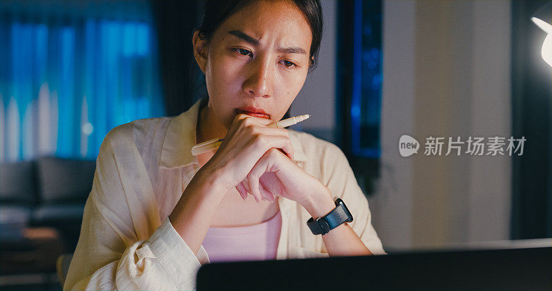 年轻的亚洲女性穿着正装衬衫坐在桌子前，拿着笔记本电脑思考和得到的想法，使紧急报告会议加班到晚上在家。