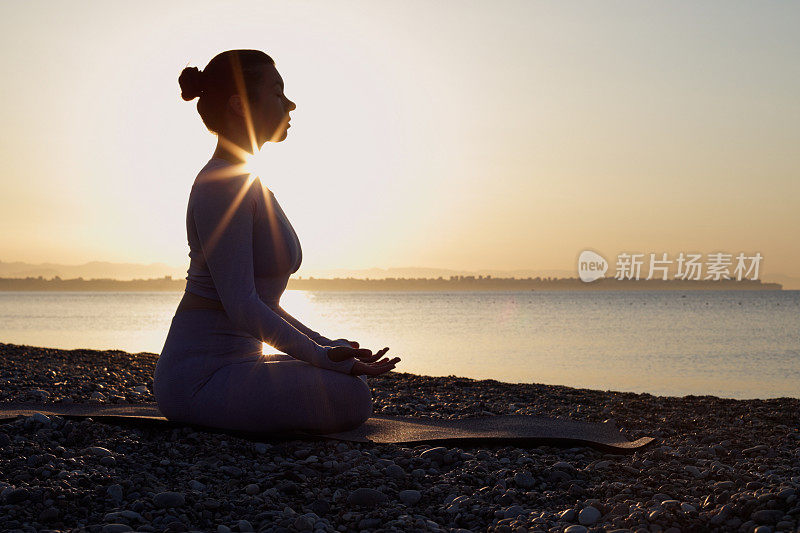 健康的年轻女子坐在瑜伽莲花姿势与闭上眼睛在大海前，在海滩上的日出冥想。