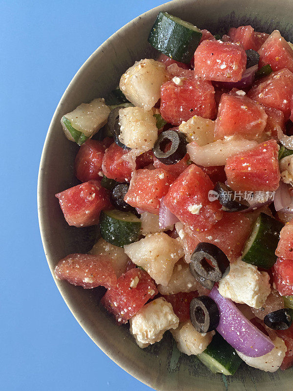 西瓜沙拉在碗的特写图像，切片黄瓜，红洋葱，羊乳酪块，薄荷叶和黑橄榄，蓝色的背景，上升的观点
