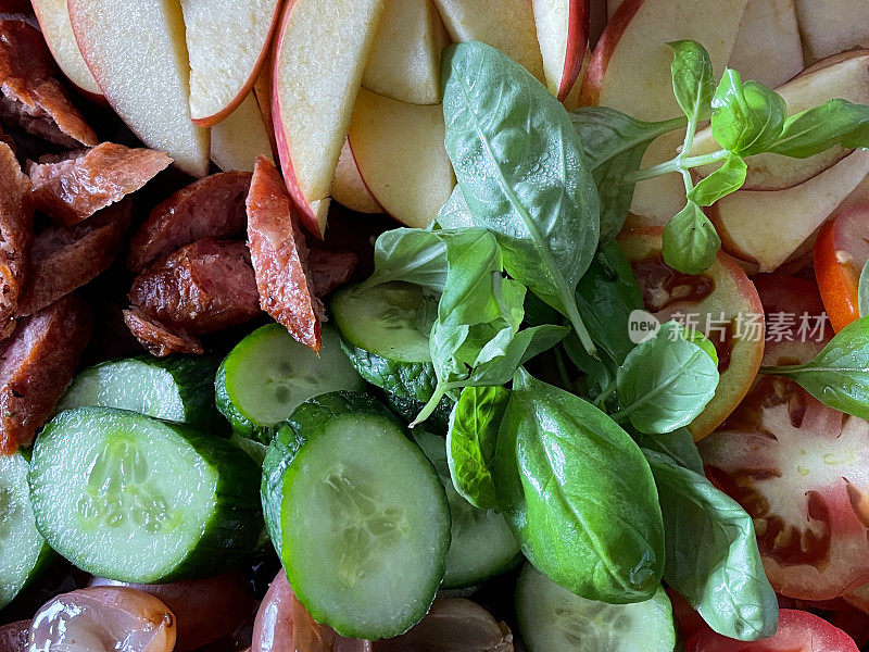 全框图像熟食板的食材包括腊肠，切碎的黄瓜，红苹果片，切片红番茄，葡萄，罗勒叶装饰，提升的食物