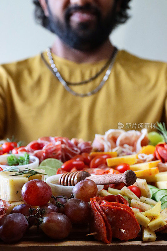 特写图像，无法识别的男子手持熟食板与蜂蜜和酸辣酱，一排排饼干，火腿和萨拉米玫瑰，面包棒，猕猴桃，藤蔓番茄，软和硬奶酪，填充橄榄，蓝莓，重点在前景