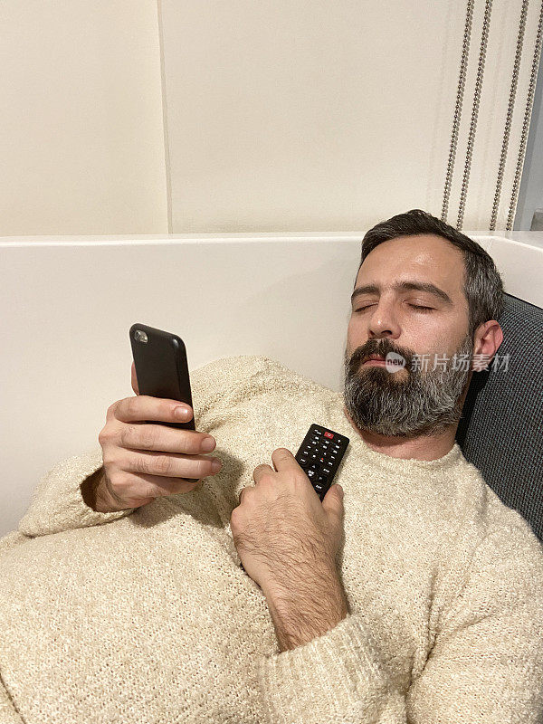 男子睡在沙发上，手里拿着智能手机和电视遥控器。睡觉的人