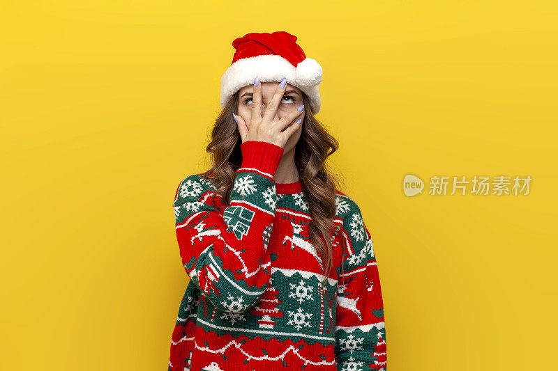 facepalm指手势。困惑的女孩穿着圣诞毛衣，戴着圣诞老人的帽子，用手捂住脸