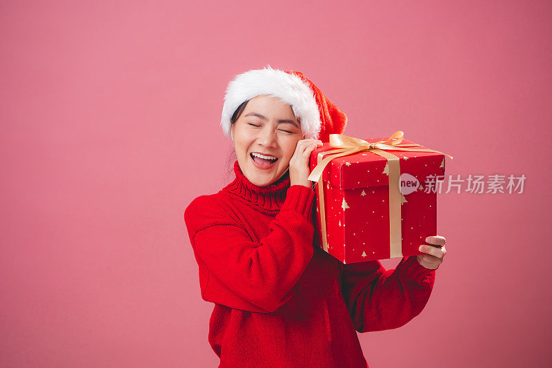 亚洲女人戴着圣诞老人的帽子，拿着礼盒开心地微笑，孤立在粉红色的背景上。