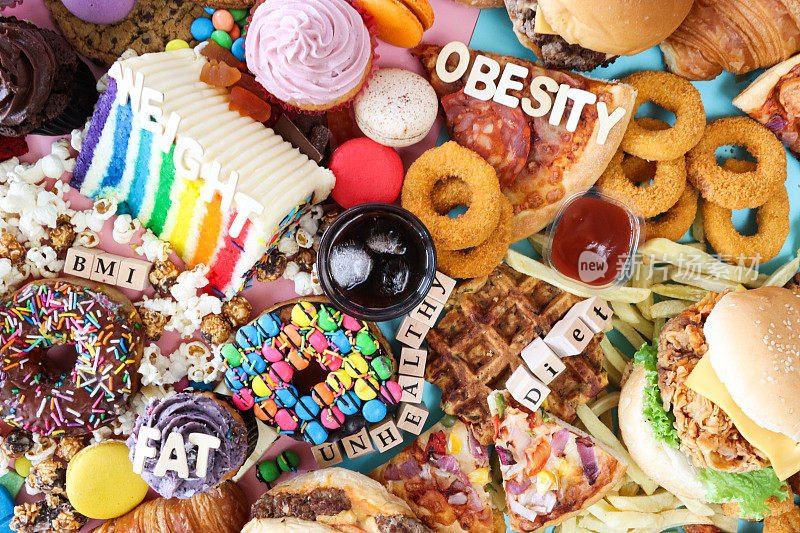 全画幅的字母骰子和木制字母拼出“脂肪”，“BMI”，“不健康”，“饮食”，“肥胖”和“体重”的甜垃圾食品，一杯冰可乐，芝士汉堡，碎洋葱圈，披萨片，土豆华夫饼，釉面圈甜甜圈