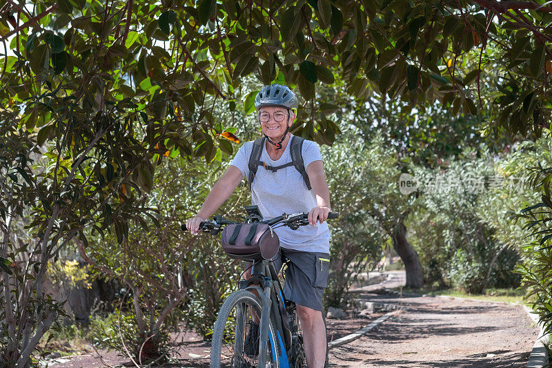 在绿色的公园里，一位带着头盔骑电动自行车的活跃微笑的老妇人。成熟迷人的女人享受退休和健康的生活方式。