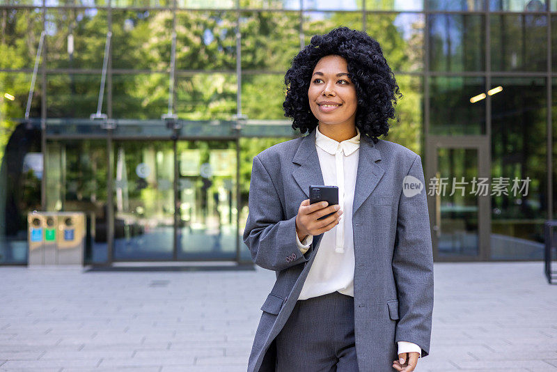 成功的非裔美国女商人从办公楼外走在城市里，金融工作者满意地微笑着，女人拿着智能手机使用应用程序，浏览社交网络