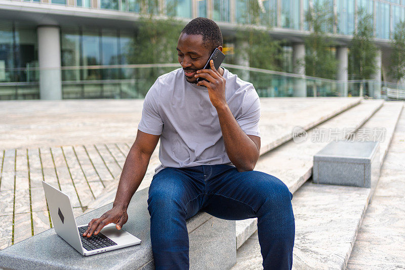 非裔美国自由职业者在城市街道上用笔记本电脑打电话。一个商人正在开虚拟会议。专业程序员使用手机和笔记本电脑在户外工作。远程工作。