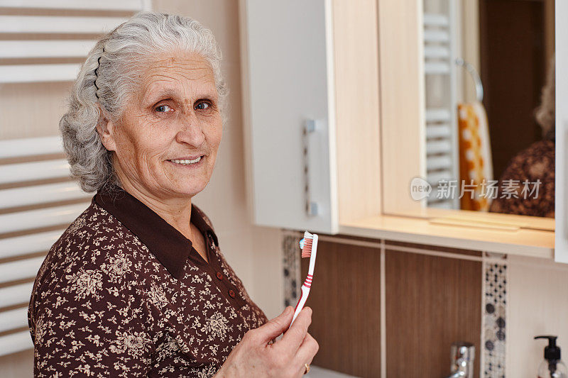 美丽的老妇人与灰色的头发拿着一个牙刷在手里，而看着镜头在浴室的镜子和水槽