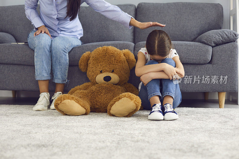 伤心的小女孩哭在地板上靠近泰迪熊，而母亲安慰她在家里。父母安慰她不幸的孩子