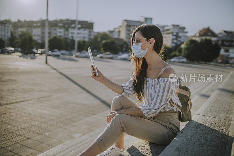 在冠状病毒大流行期间，戴医用口罩的妇女在城市使用电子平板电脑，坐在楼梯上