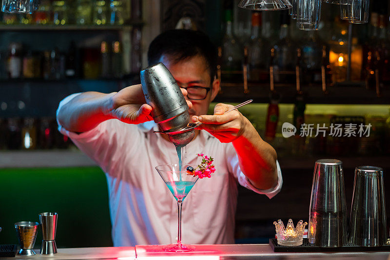 专业的亚洲调酒师，在夜总会吧台将混合酒倒入装饰好的鸡尾酒杯中，为顾客服务。男调酒师调酒师准备酒精饮料与烈酒架子的背景