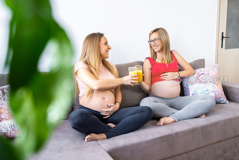 两个开心的孕妇坐在沙发上喝着新鲜的橙汁