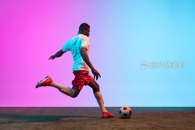 年轻的非洲男子，职业足球运动员踢足球在草地地板上孤立的梯度蓝色粉红色的背景