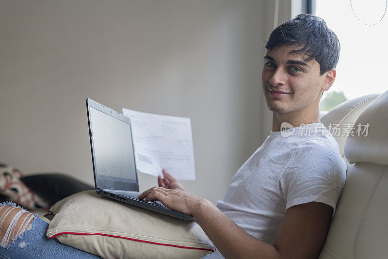 一个少年坐在客厅的沙发上，一边用笔记本电脑做作业，一边阅读他必须完成的文件