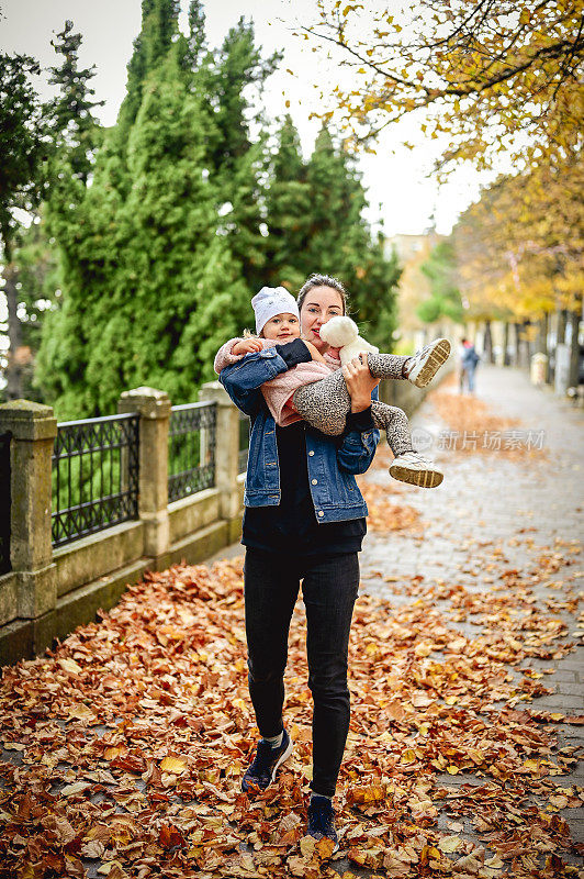 一位年轻的母亲和她的女儿在秋天的公园里玩耍。在满是黄叶的街道上，妈妈把女孩抱在怀里。