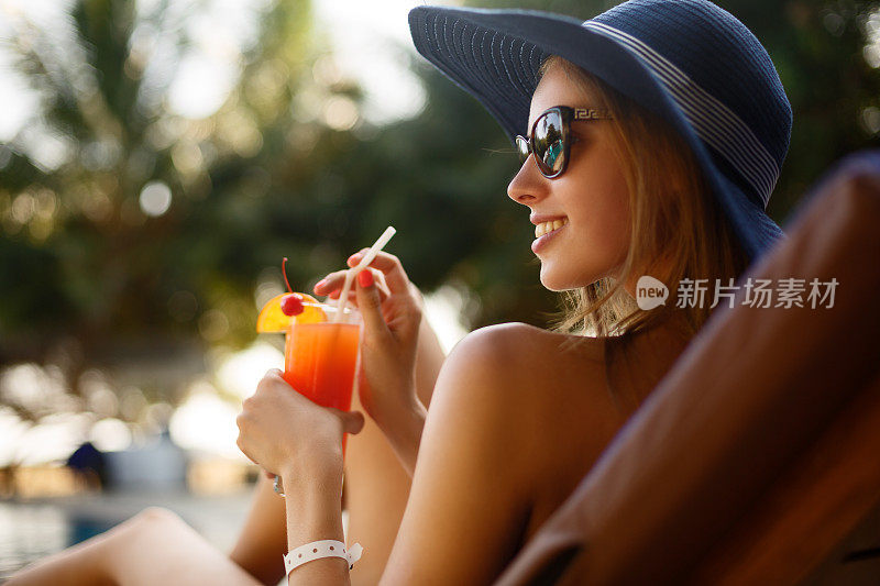 一个年轻女人的肖像与鸡尾酒杯在热带阳光下寒冷在游泳池附近的躺椅上，后面有棕榈树。假期的概念