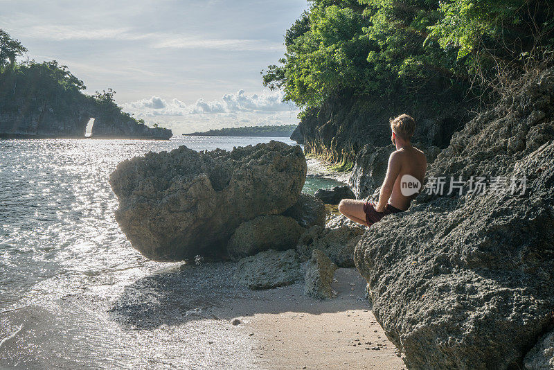 年轻人沉思在岩石上的热带海滩在巴厘岛享受海滩假日在热带气候的目的地-人们旅行度假海滩概念