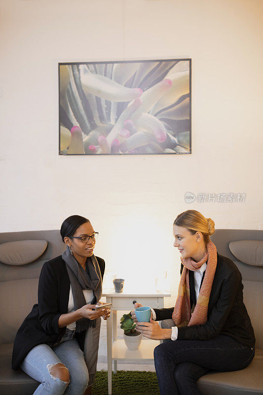 两位年轻女性坐在客厅里，一边玩着触屏智能手机，一边浏览互联网，一边聊天，喝着咖啡。