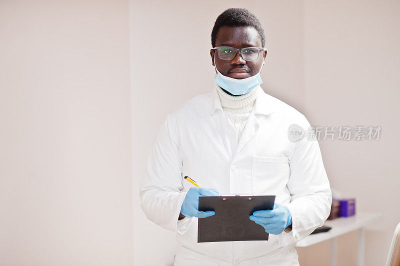 戴眼镜和面具的非裔美国男医生手持黑色写字板在诊所里摆姿势。