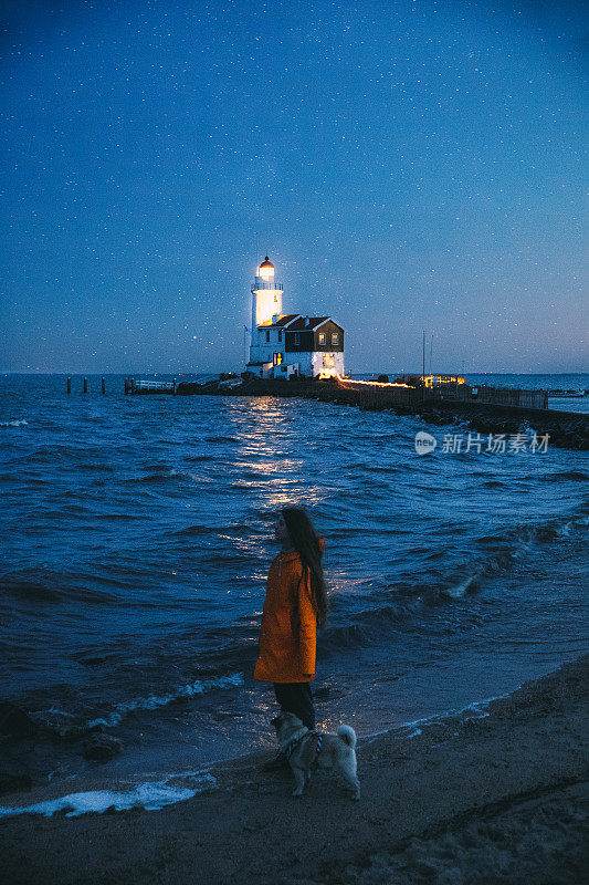 在荷兰，一个穿着雨衣的女人带着她的狗在美丽的灯塔附近的海滩上散步，欣赏星空