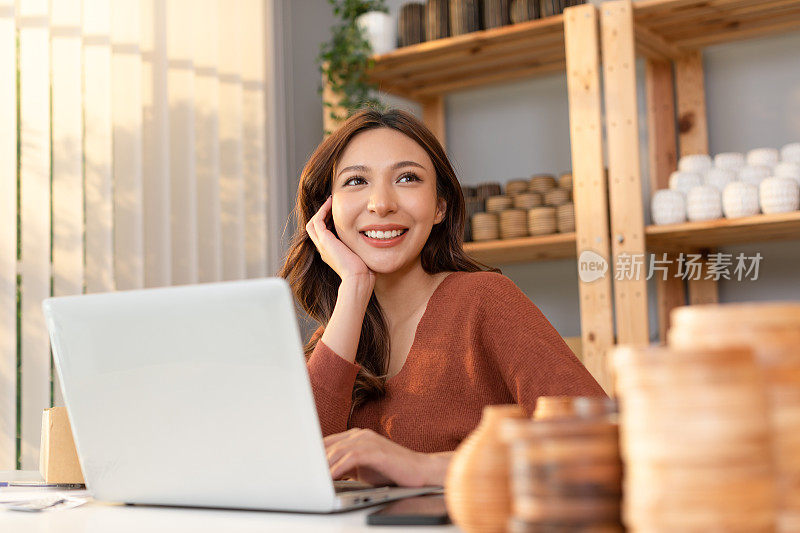 快乐的亚洲年轻企业主花瓶店工作和网上销售电脑笔记本电脑微笑，抬头。自信的女企业家在家里被美丽的花瓶包围