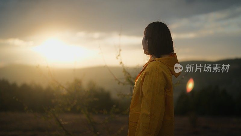 一名女子在寒冷多雨的天气里穿着黄色雨衣，在山上看大自然中的太阳