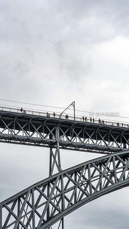 游客们在唐路易斯一号钢桥的上层平台上散步，拍照，一名女子在那里摆姿势，波尔图地铁在雨云中穿行。