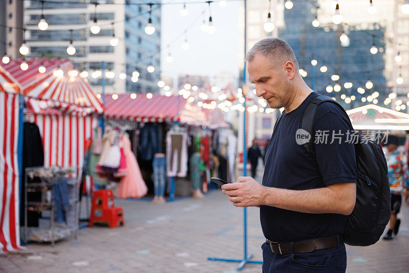 一个背着背包的人站在灯火通明的城市街市上，用着手机