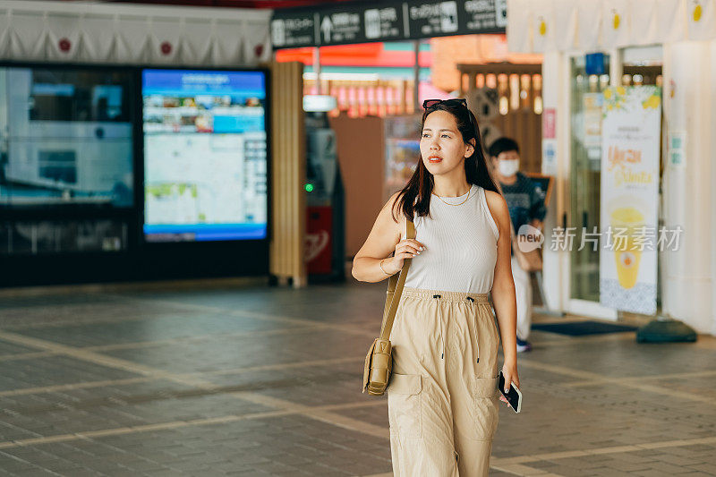 这位亚洲女性自信地大步走进室内，目光投向前方，周围是商店和充满活力的招牌。气氛很轻松，她的表情流露出一种轻松和满足的感觉。