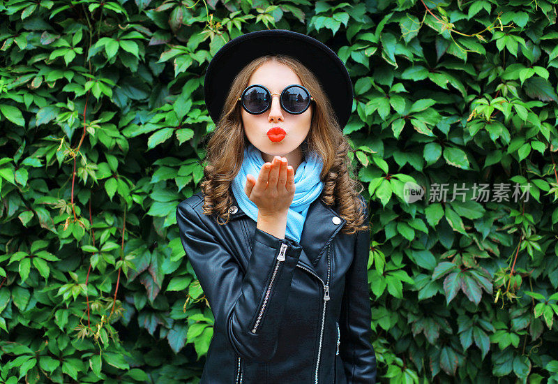 时尚肖像年轻女子送飞吻吹红唇戴黑帽子在绿色树叶的背景