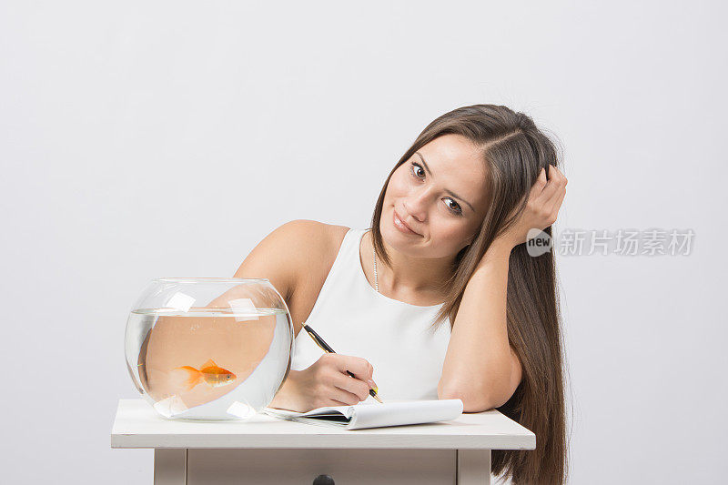 女孩在笔记本上写字，站在鱼缸旁边养金鱼