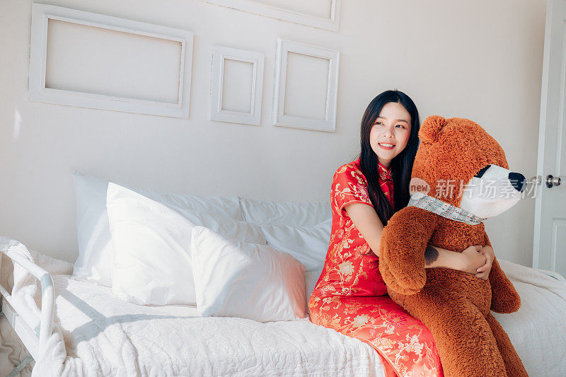 亚洲农历新年妇女的礼物旗袍，旗袍拥抱熊坐在床边靠近窗户在干净的白色房间。