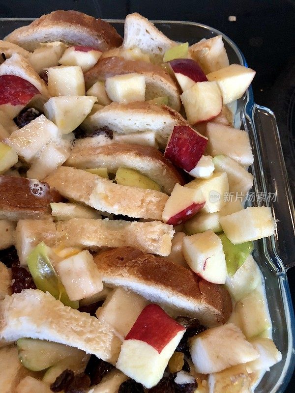 这是一个玻璃烤盘，里面装着准备好的多层面包和黄油布丁的原料，涂了黄油的白面包片，上面撒着干果和红绿苹果丁