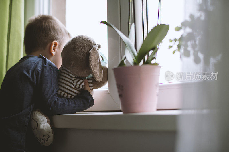 小男孩戴着防护口罩，带着闷热的玩具，从窗户往外看。预防传染病