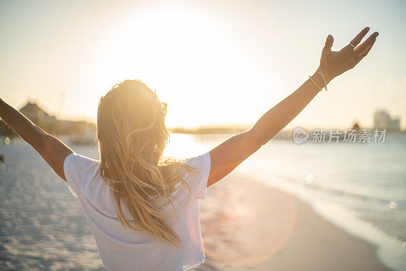 夕阳西下，拥抱大自然的快乐年轻女子;女人站在沙滩上，伸开双臂