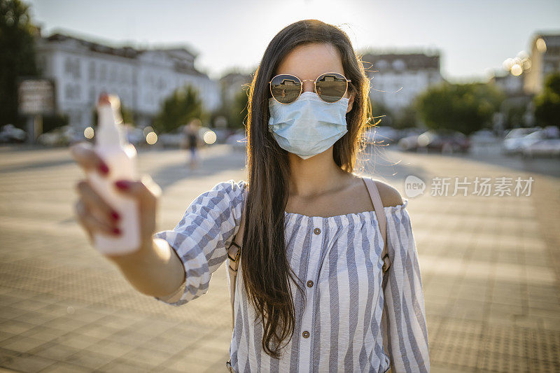 一名年轻女子戴着外科口罩，手上涂着洗手液，在封锁期间站在空荡荡的城镇广场上