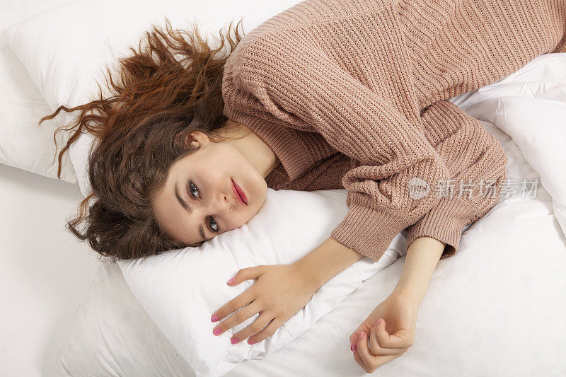 年轻女子躺在床上。她穿着一件米色毛衣，看着镜头。