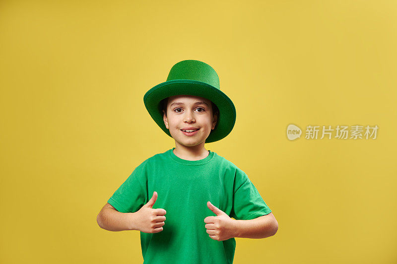 微笑的小男孩在爱尔兰绿色妖精帽子显示竖起的大拇指站在黄色背景与拷贝空间。圣帕特里克日