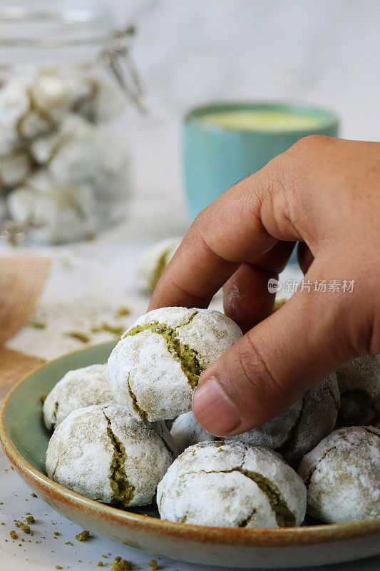 不认识的人选择个人的图像，新鲜出炉的抹茶苦杏仁饼干从盘子旁边有盖的玻璃饼干罐和马克杯，碎屑在大理石效果的背景，关注前景