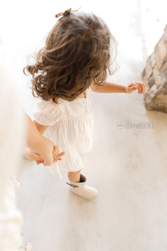 一个三岁的小女孩穿着白色的裙子，棕色的蝴蝶结和白色的雨靴，牵着妈妈的手走在白色的乡间门廊上