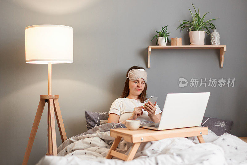 平静微笑的女人戴着睡眠面具坐在床上，拿着笔记本电脑，拿着手机，查看社交网络或邮箱，享受早上的空闲时间。