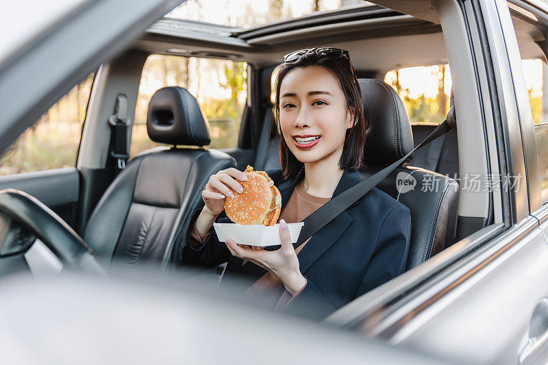 亚洲女人在车里休息，吃汉堡。一个年轻成功快乐的女人正在开车。