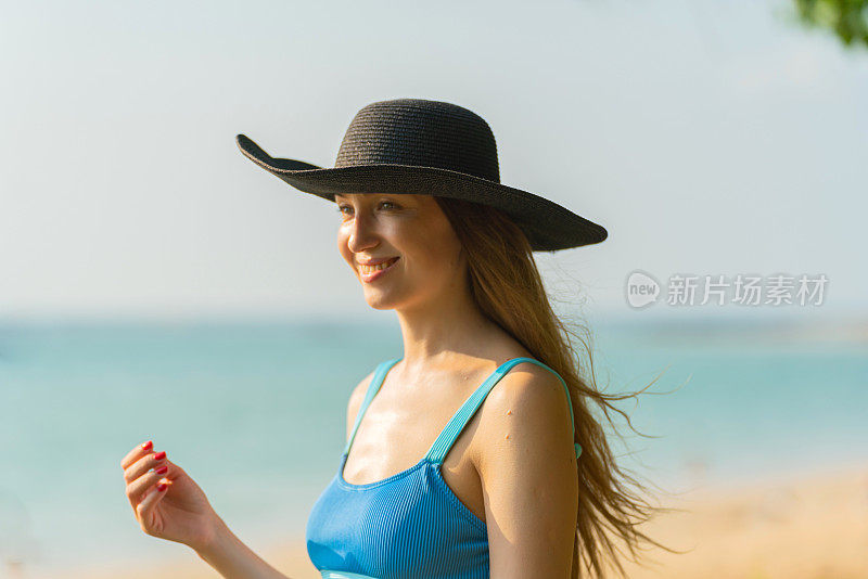 生活是一片海滩。美丽的白人女子在蓝色比基尼黑色沙滩帽和太阳镜漫步在海滩上享受海风和太阳晒黑