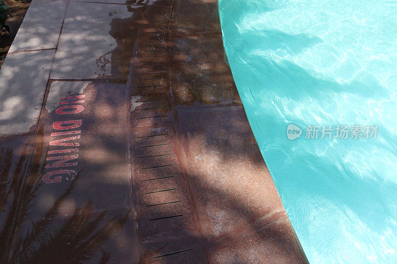 室外特写，游泳池碧蓝的马赛克瓷砖被荡漾的水扭曲，泳池边的石头露台方板上画着禁止潜水的警告标志，溅起的水，高架的景色