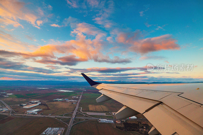 飞机在夕阳的天空中飞过城市和飞机的机翼。从飞机窗口看到的景象。在空中旅行。