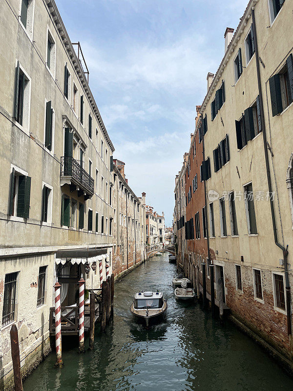 Cofano(小型舷外摩托艇)在意大利威尼斯狭窄的运河中航行的特写图像，红白条纹系泊杆，横跨桥梁，多云的蓝色晴朗的天空，小的缩小视角，聚焦于前景