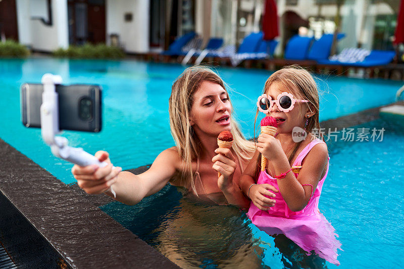 母女俩在泳池里享用冰淇淋，手里拿着自拍杆，在碧水边捕捉笑容。穿着泳装的妈妈和女孩分享甜蜜的时刻，用智能手机记录夏天的回忆。
